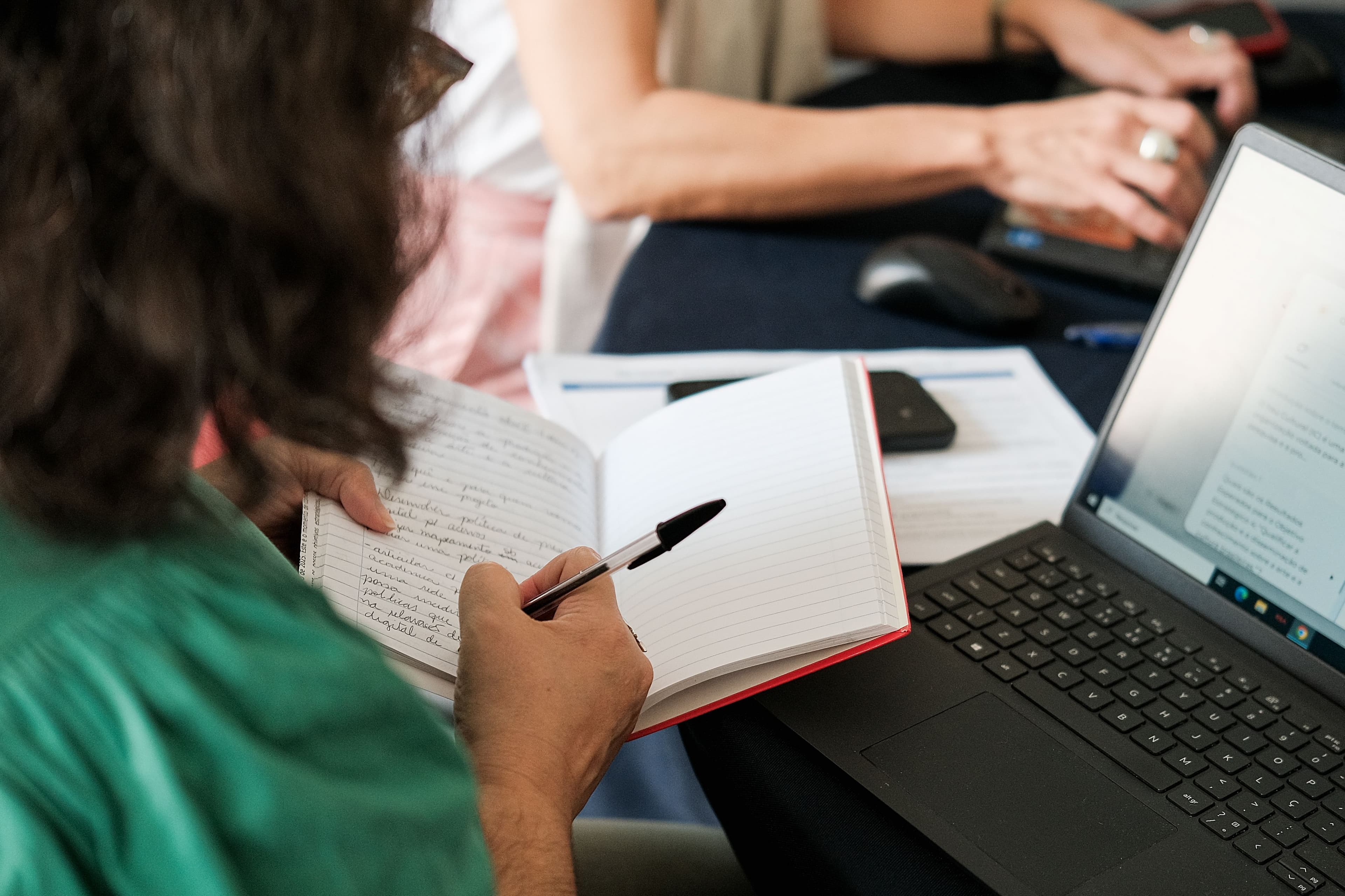 Uma mulher de costas escrevendo em um caderno, em frente a um laptop e ao fundo se vê as mãos de outra mulher digitando